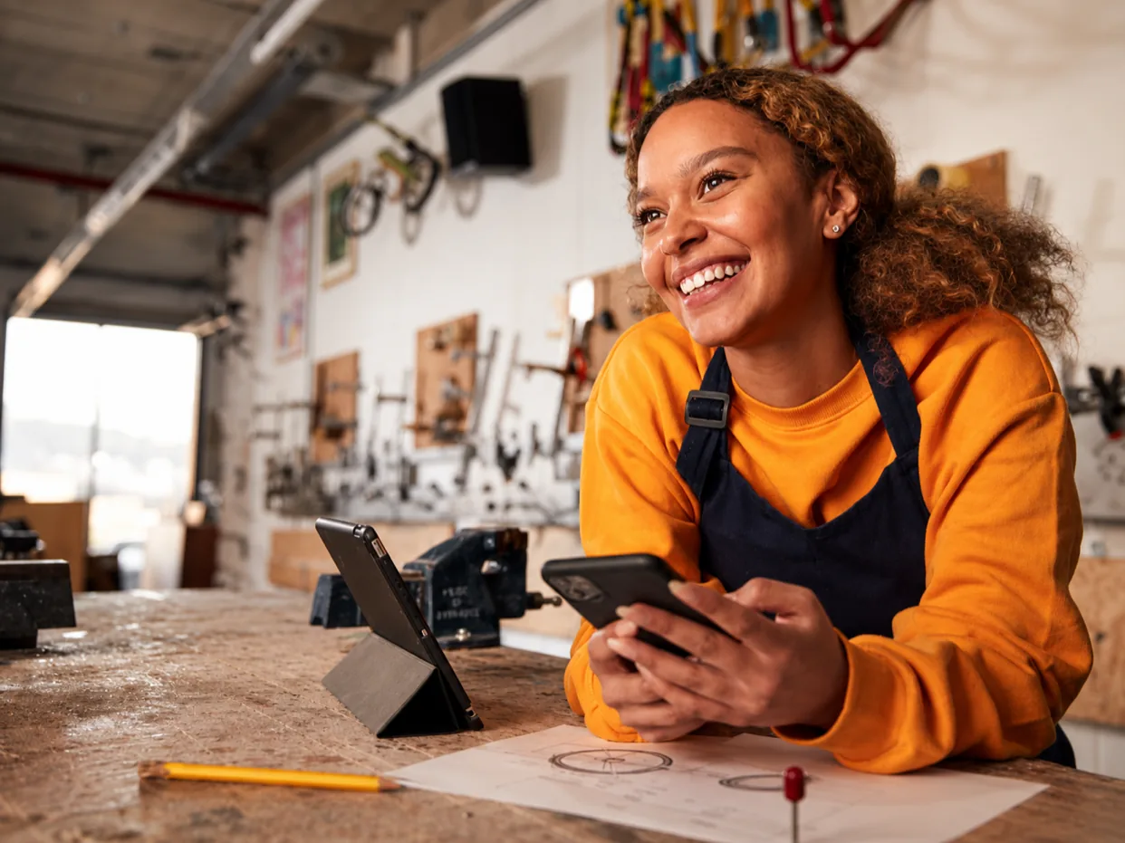 A technician in a repair workshop holding a phone, a tablet on the bench beside her.